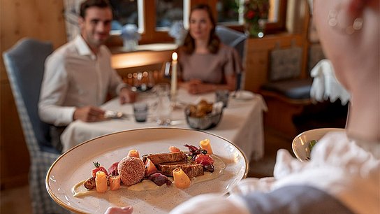 Kellnerin serviert Gourmet Menü, im Hintergrund ist ein Pärchen beim Candlelight Dinner zu sehen
